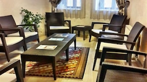 Une pièce avec un plancher en bois, une table basse au centre et plusieurs chaises disposées autour. On aperçoit, à l’arrière-plan, une plante en pot et une fenêtre avec des rideaux.