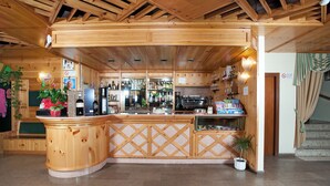 A wooden bar area with a counter, shelves with bottles, and a kitchen in the background. - Magnola Palace Hotel (Ovindoli)