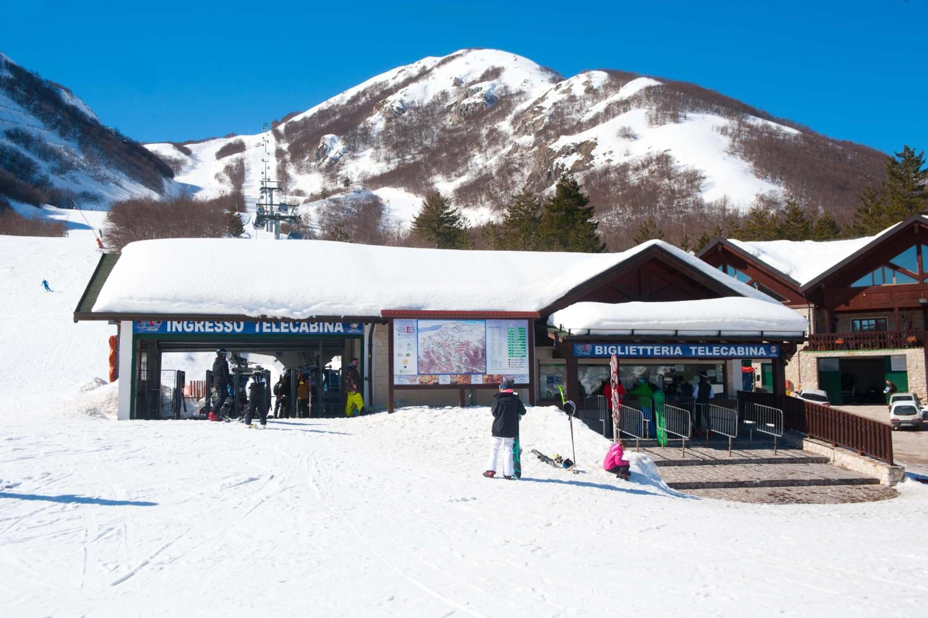 A ski resort with a telecabina, a mountain backdrop, and a building with a sign reading "INGRESSO TELECABINA".