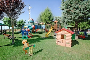 A playground with various play structures including slides and climbing areas, surrounded by trees and a bench.