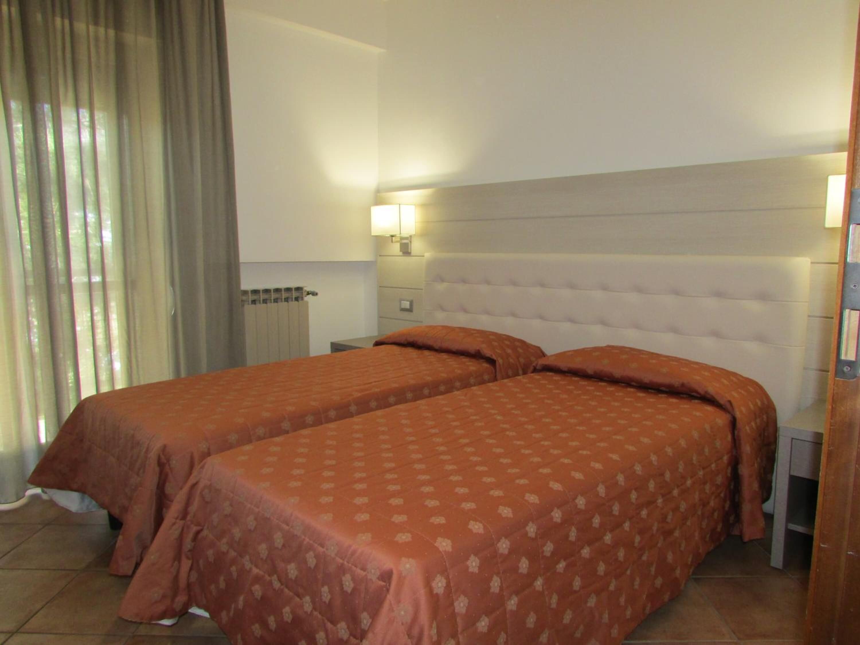 A hotel room with two beds, a headboard, a nightstand, a lamp, and a window with curtains.