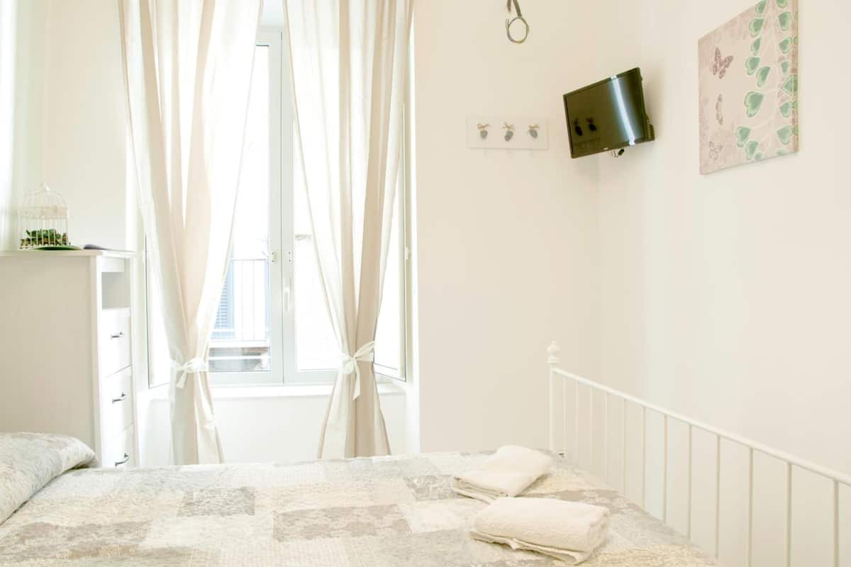 A bedroom with a bed, a window with curtains, a wall-mounted light, and a small shelf with a decorative item.
