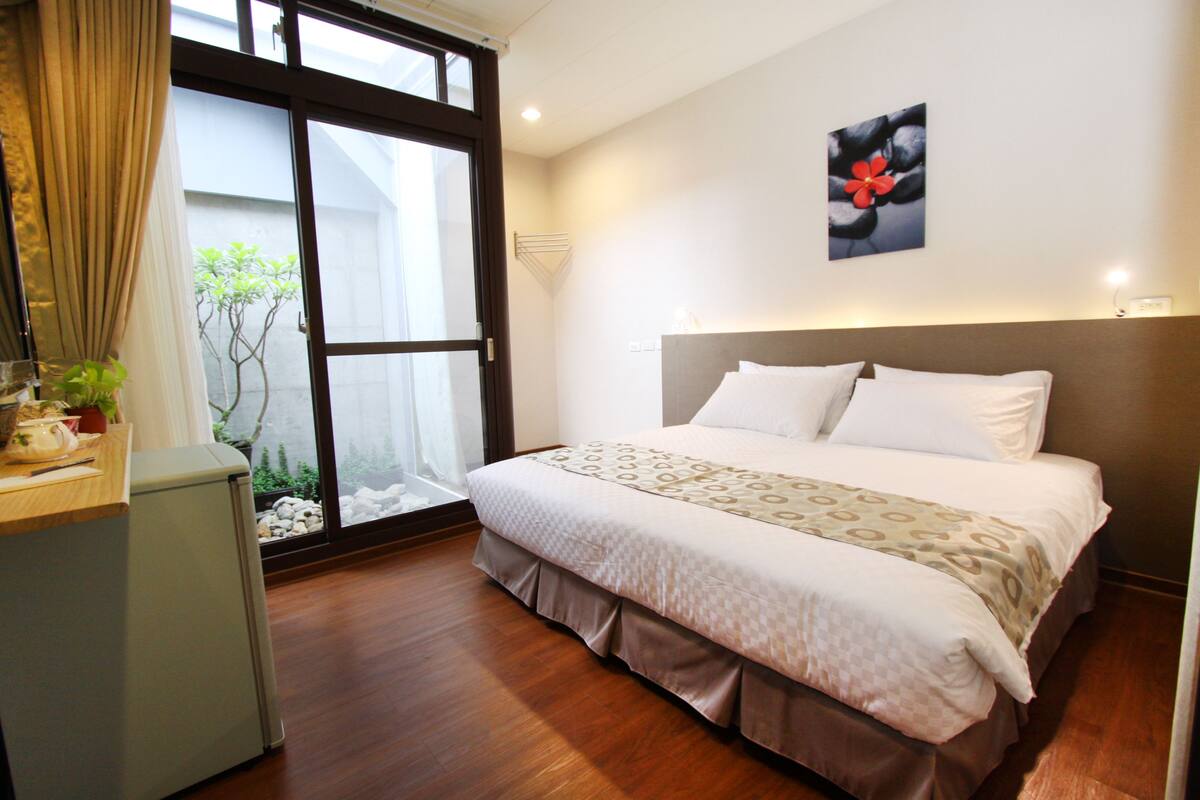 A hotel room with a bed, a desk, and a view of a garden through a sliding door.
