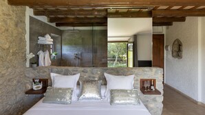 Une chambre avec un mur en pierre, un grand lit, un plafond en bois, une salle de bain avec une douche en verre et une vue sur l’extérieur.
