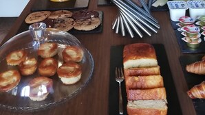 Un coin petit-déjeuner avec des fruits, des viennoiseries et une carafe de jus, installé dans une pièce rustique aux murs de pierre, avec un lit et un espace salon.