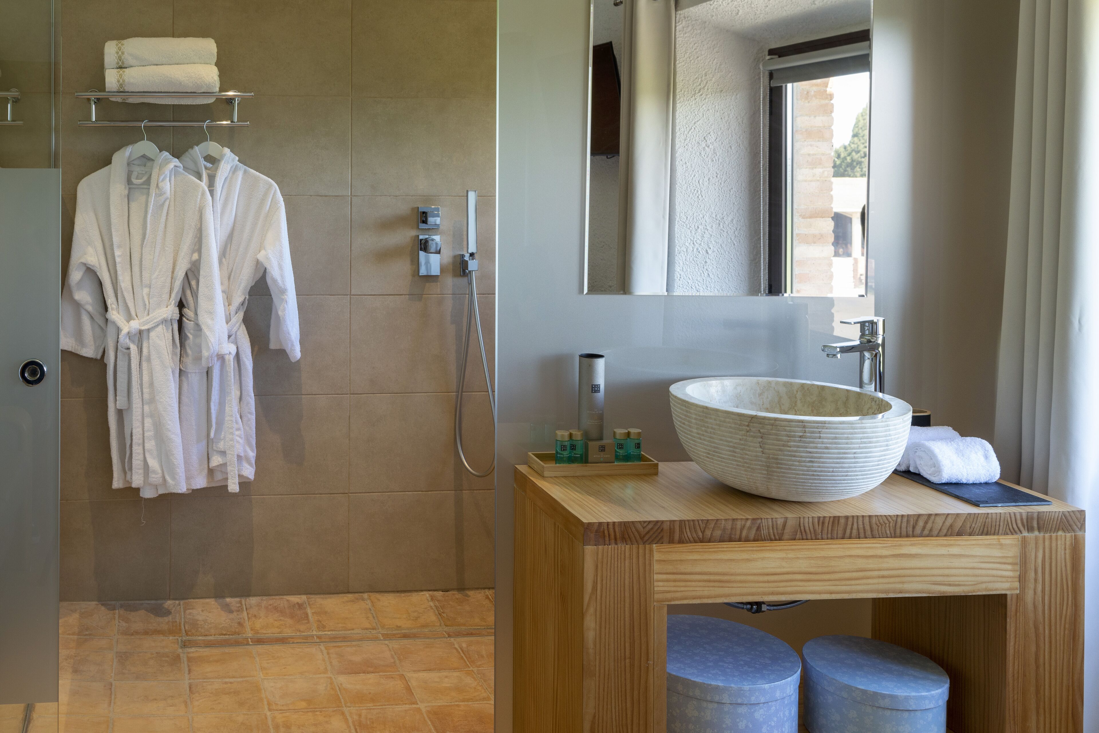 Un baño con un espejo grande, un tocador de madera, un lavabo grande tipo cuenco y dos taburetes azules. Hay dos batas blancas colgadas en un perchero.