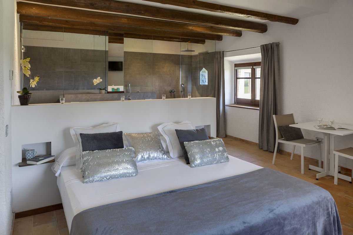 Quarto com cama grande, mesa de cabeceira, espelho, janela pequena e vista para o banheiro.
