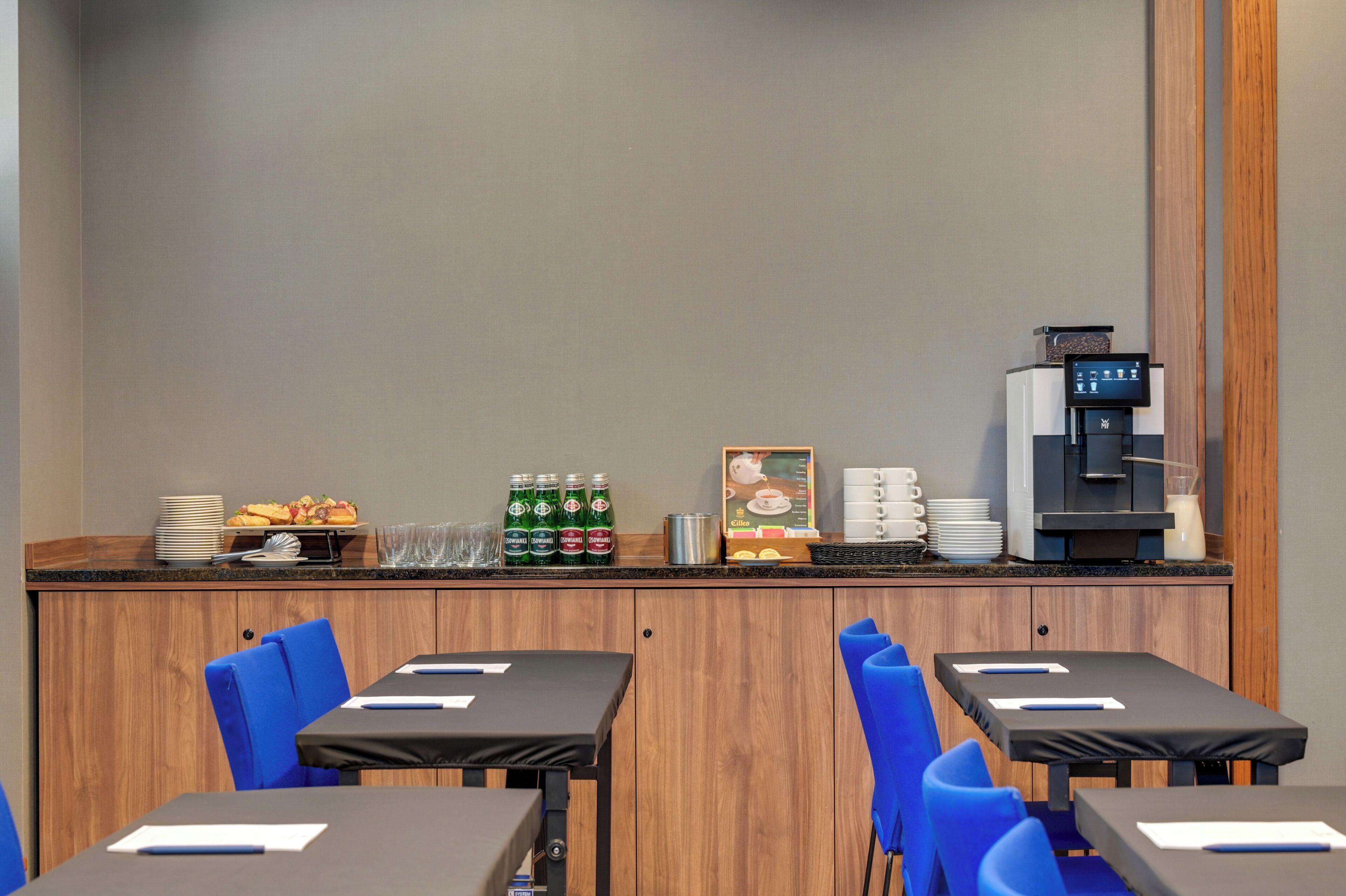 Uma mesa de pequeno-almoço com pratos, chávenas e uma máquina de café. Há cadeiras e mesas azuis em primeiro plano.