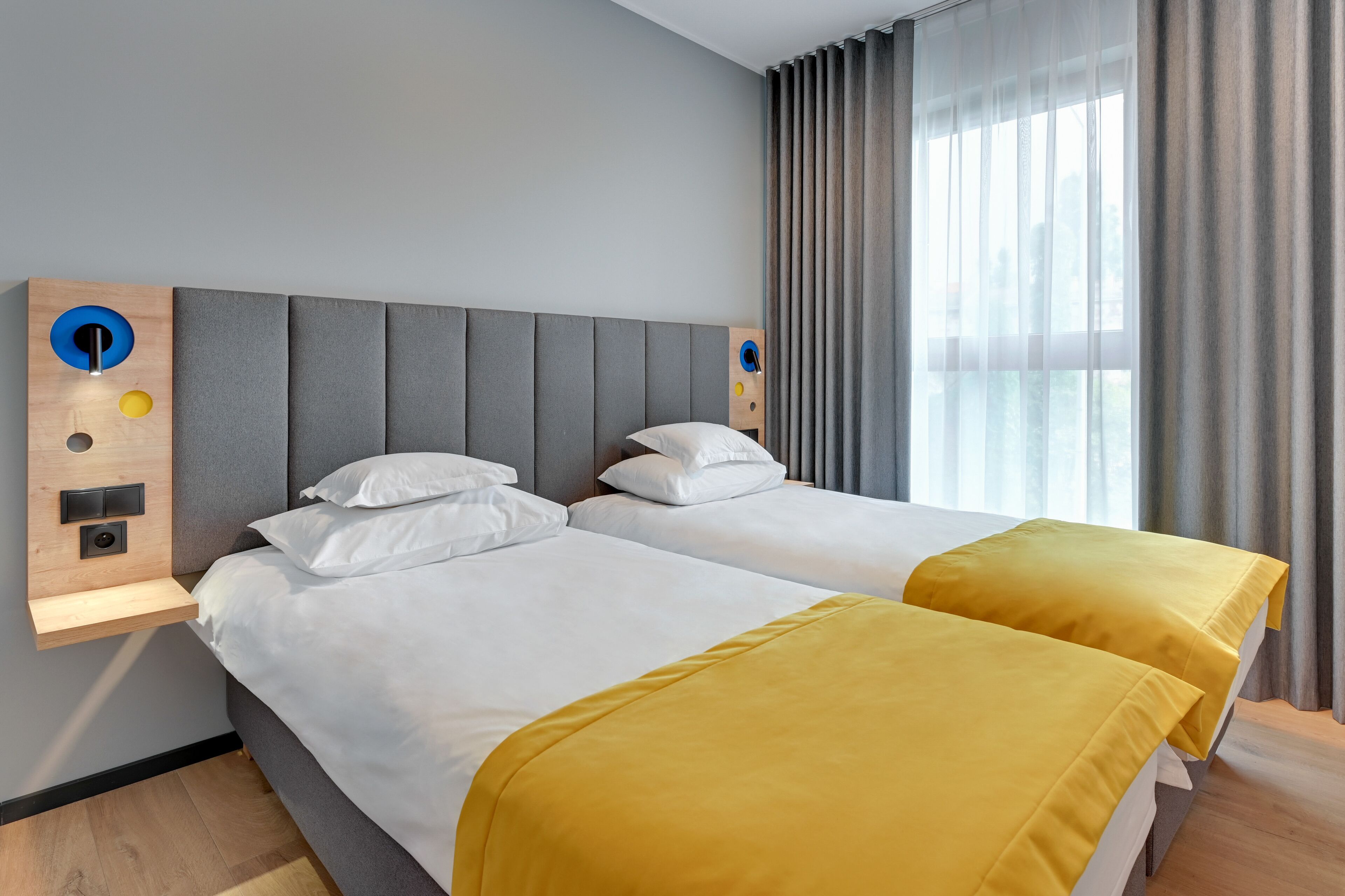 A hotel room with two beds, a wooden headboard, a small wooden shelf with a light fixture, and a large window with curtains.