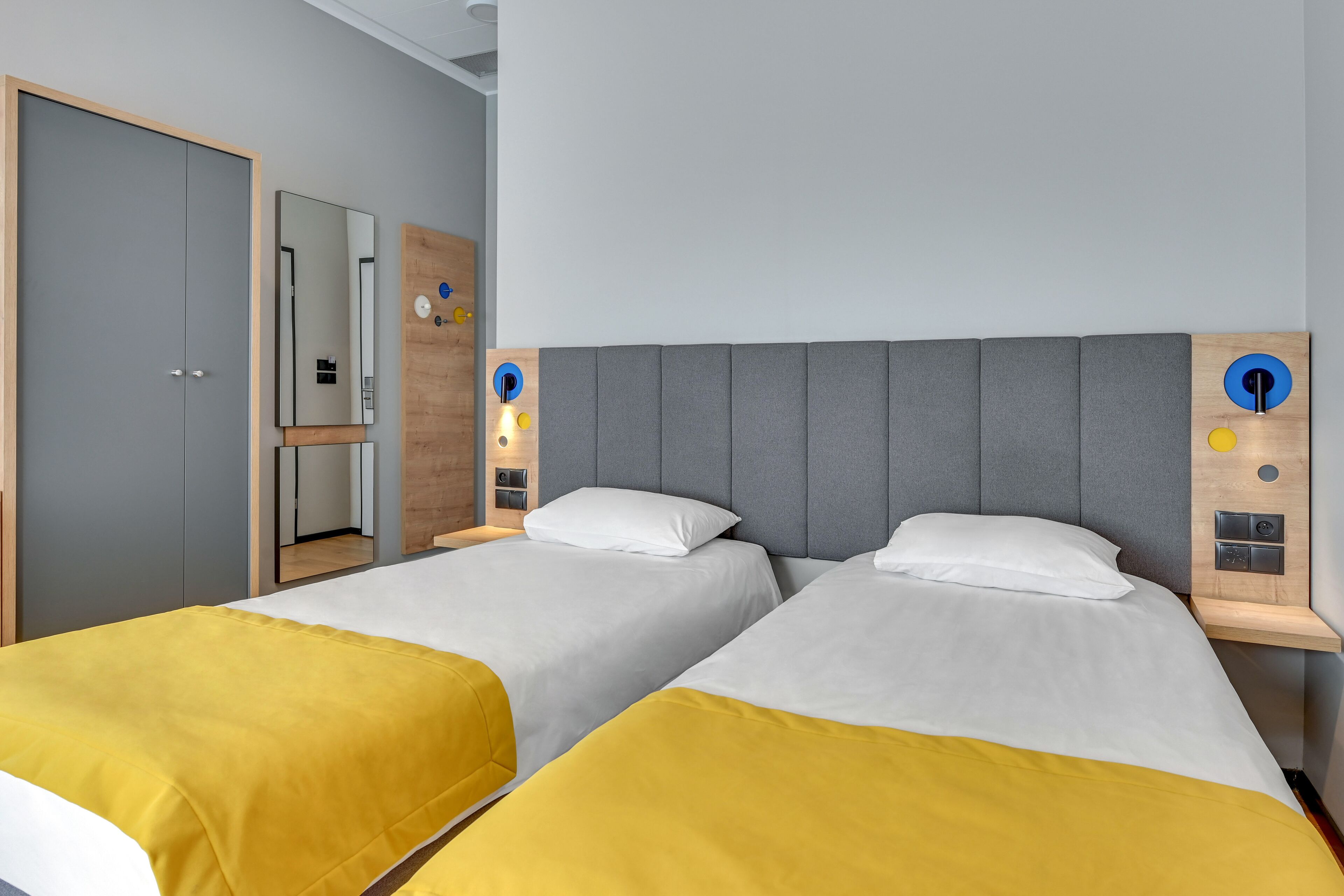A hotel room with two beds, each with a white and yellow bedspread, a grey upholstered headboard, and a wooden nightstand with a mirror and wall-mounted lights.