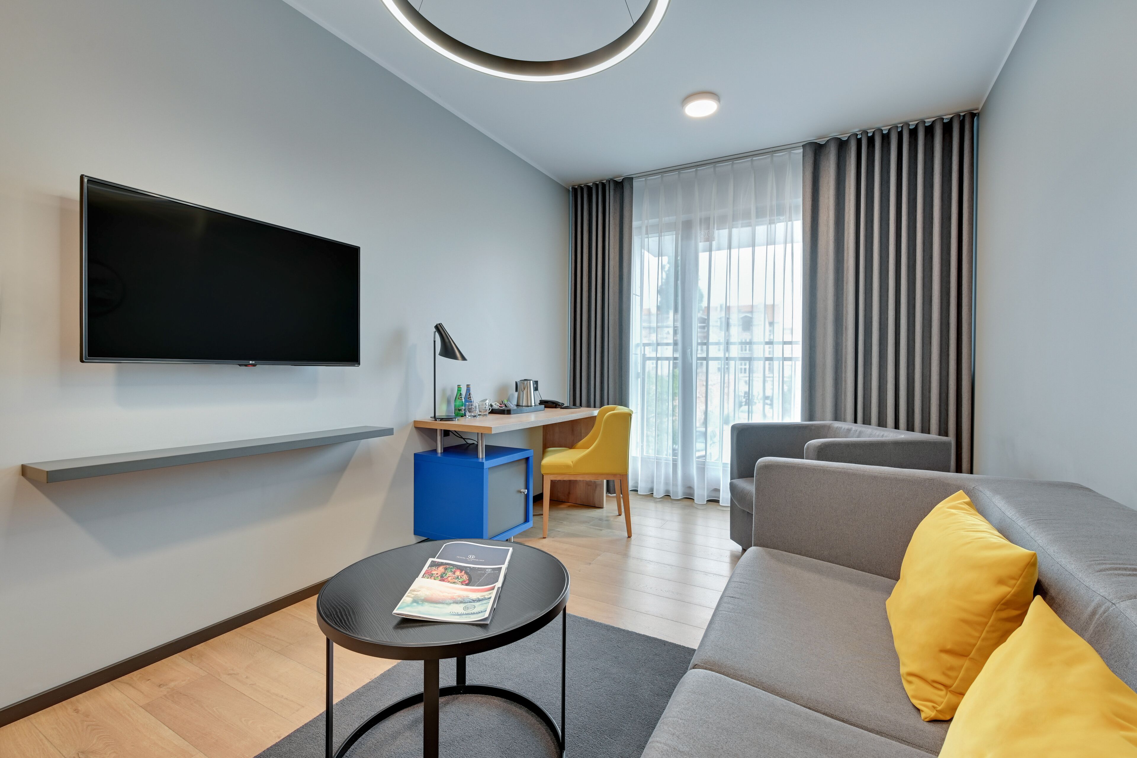 A modern living room with a flat-screen TV, a sofa with yellow cushions, a small round table, and a desk with a chair.