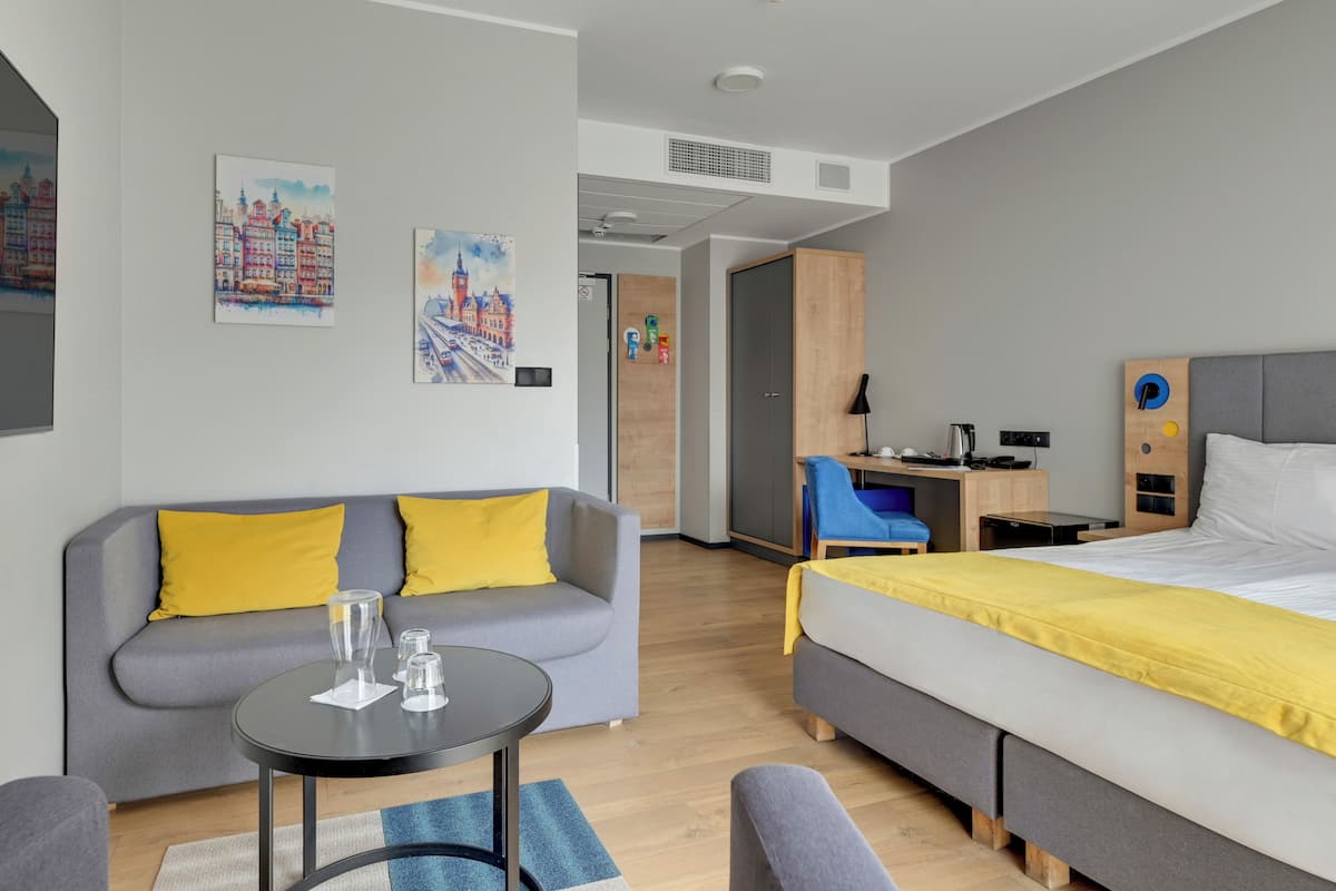A modern hotel room with a grey sofa, a bed with a yellow bedspread, a desk with a chair, and artwork on the walls.