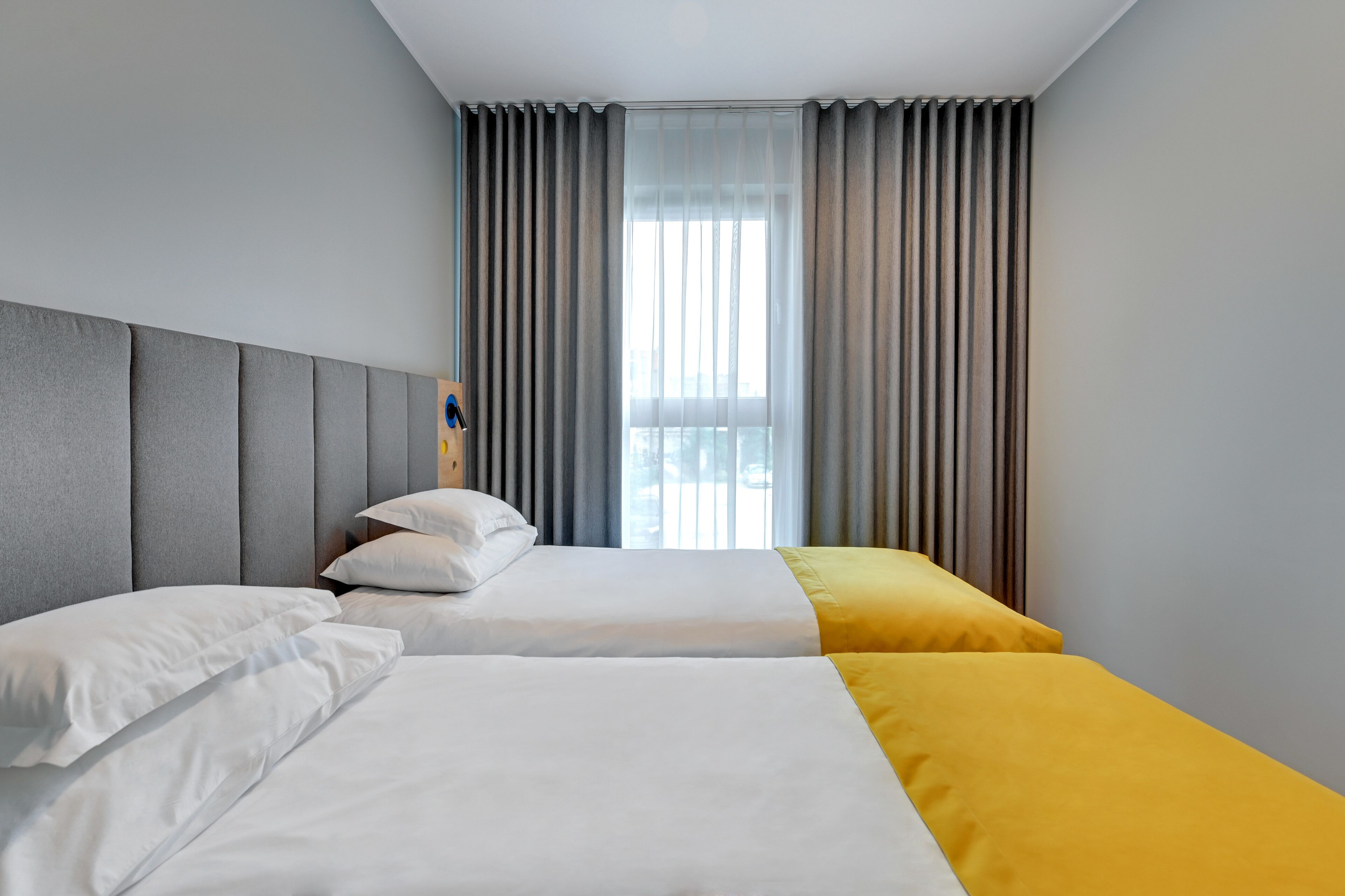 Habitación de hotel con dos camas, cabecera gris, ropa de cama blanca y amarilla, y una ventana con cortinas grises.