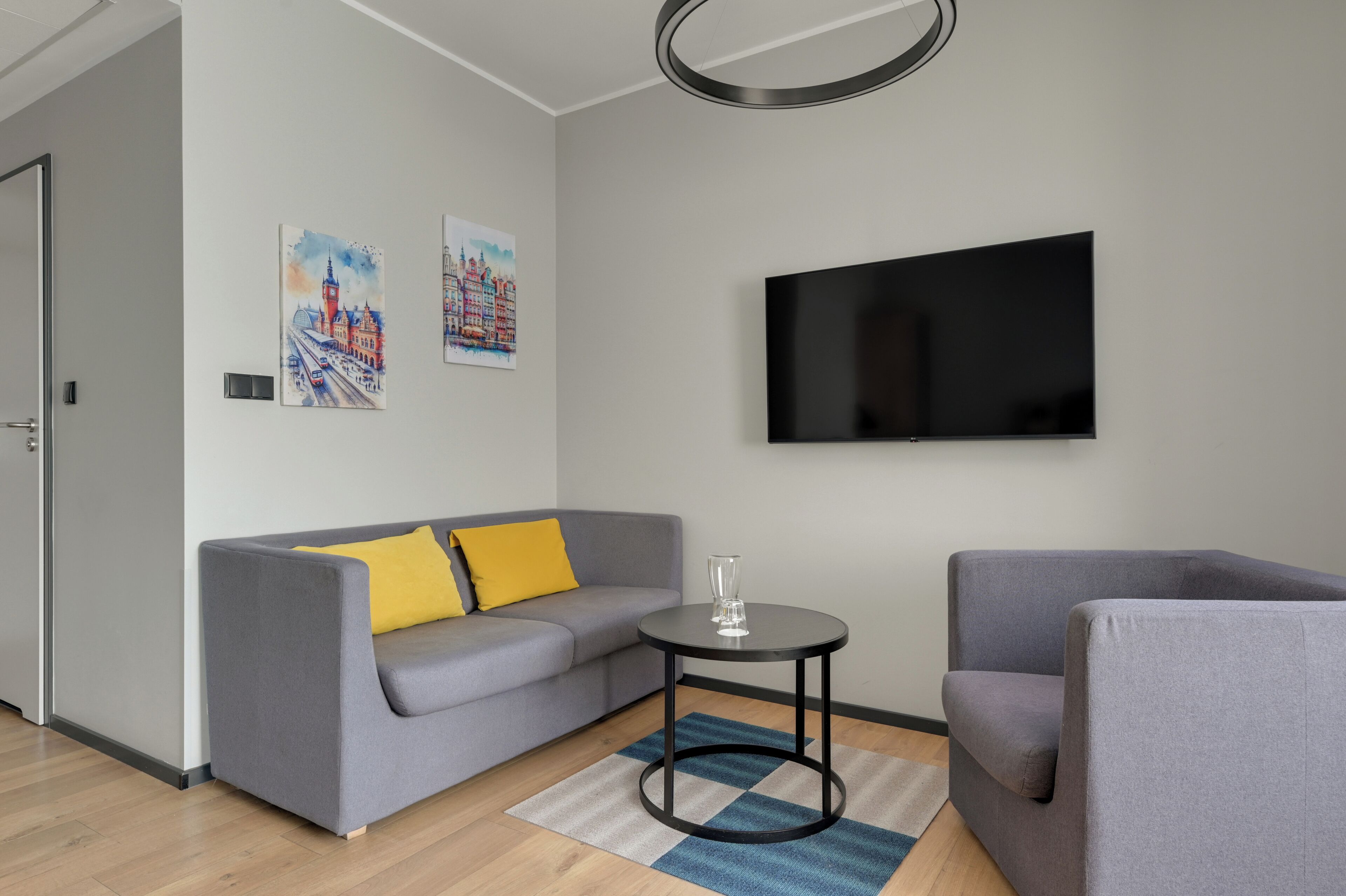 A modern living room with a grey sofa, a small round table, a flat-screen TV, and two paintings on the wall.