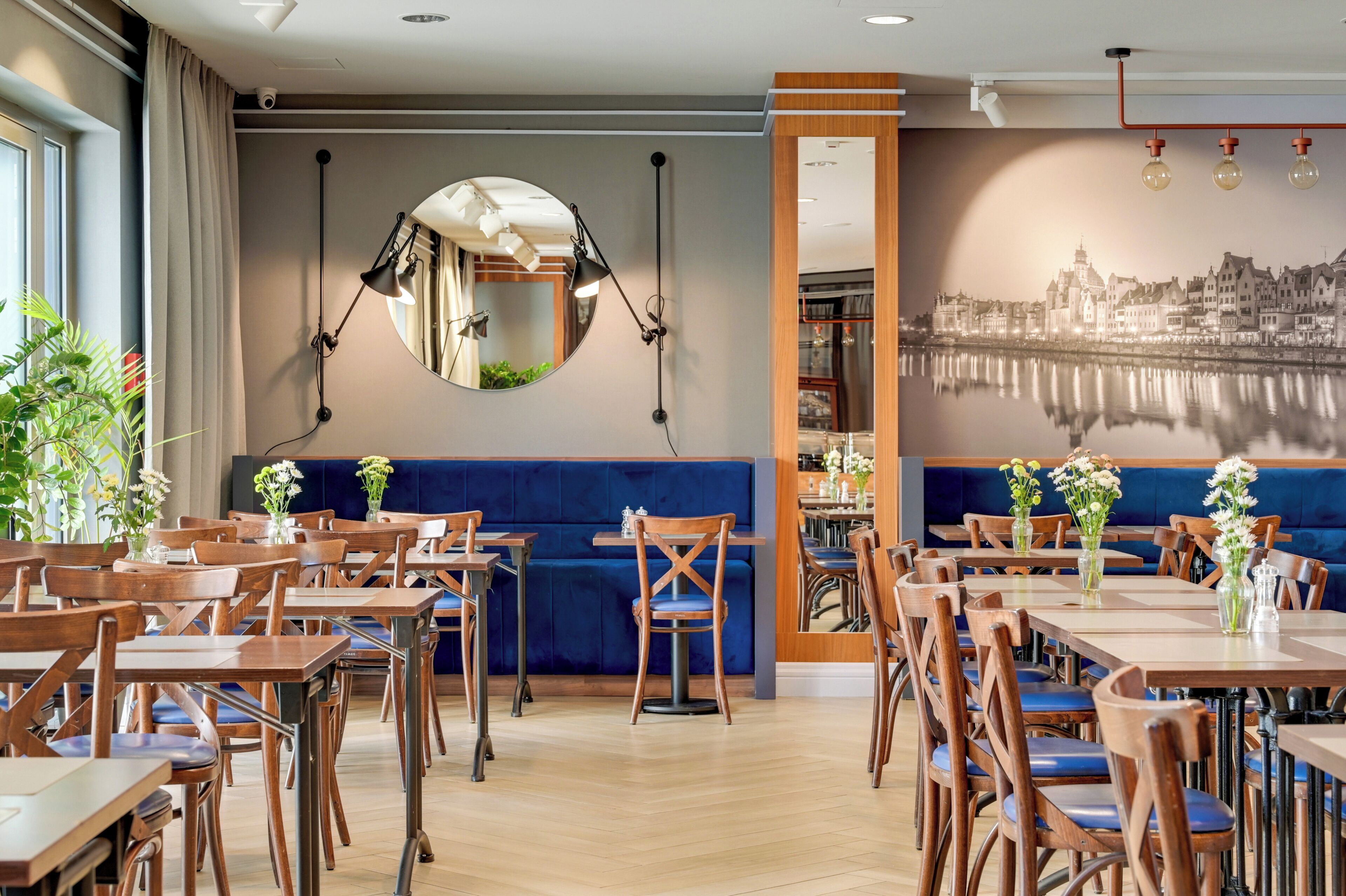 A restaurant with wooden tables and chairs, a blue cushioned bench, and a cityscape mural on the wall.