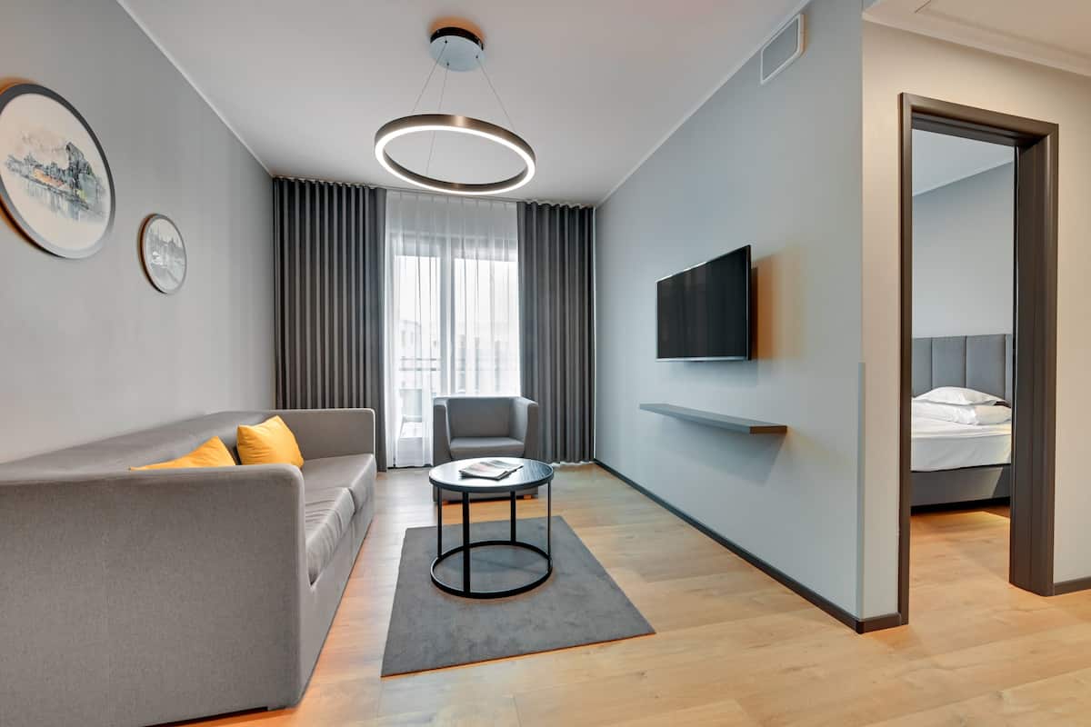 A modern hotel room with a grey sofa, a round coffee table, a TV mounted on the wall, and a bed with a grey headboard.