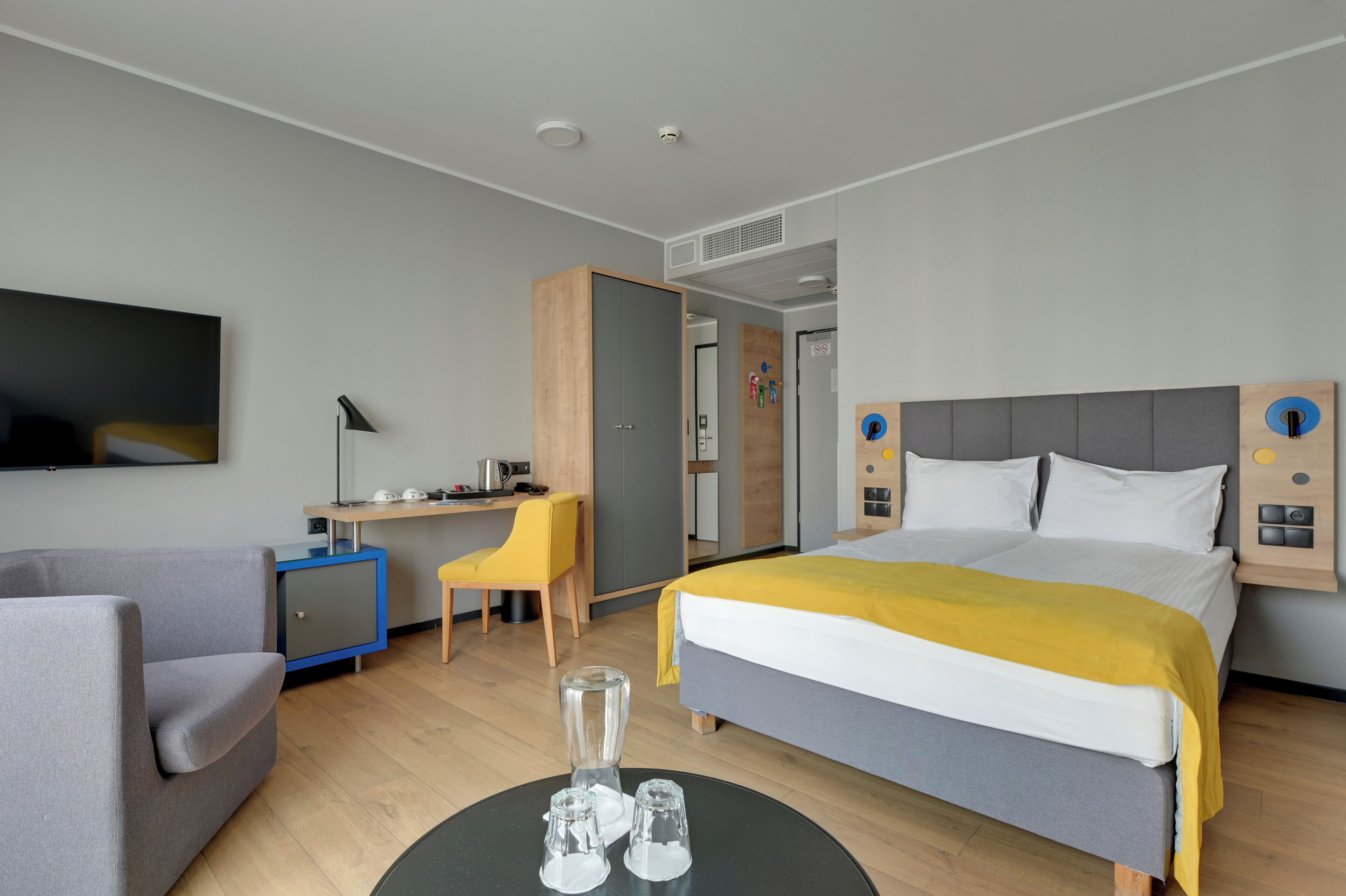 A modern hotel room with a bed, a desk, a chair, a TV, and a wardrobe.