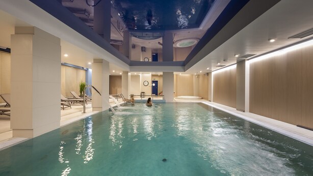 Una piscina interior con agua cristalina, techo reflectante y un reloj en la pared.
