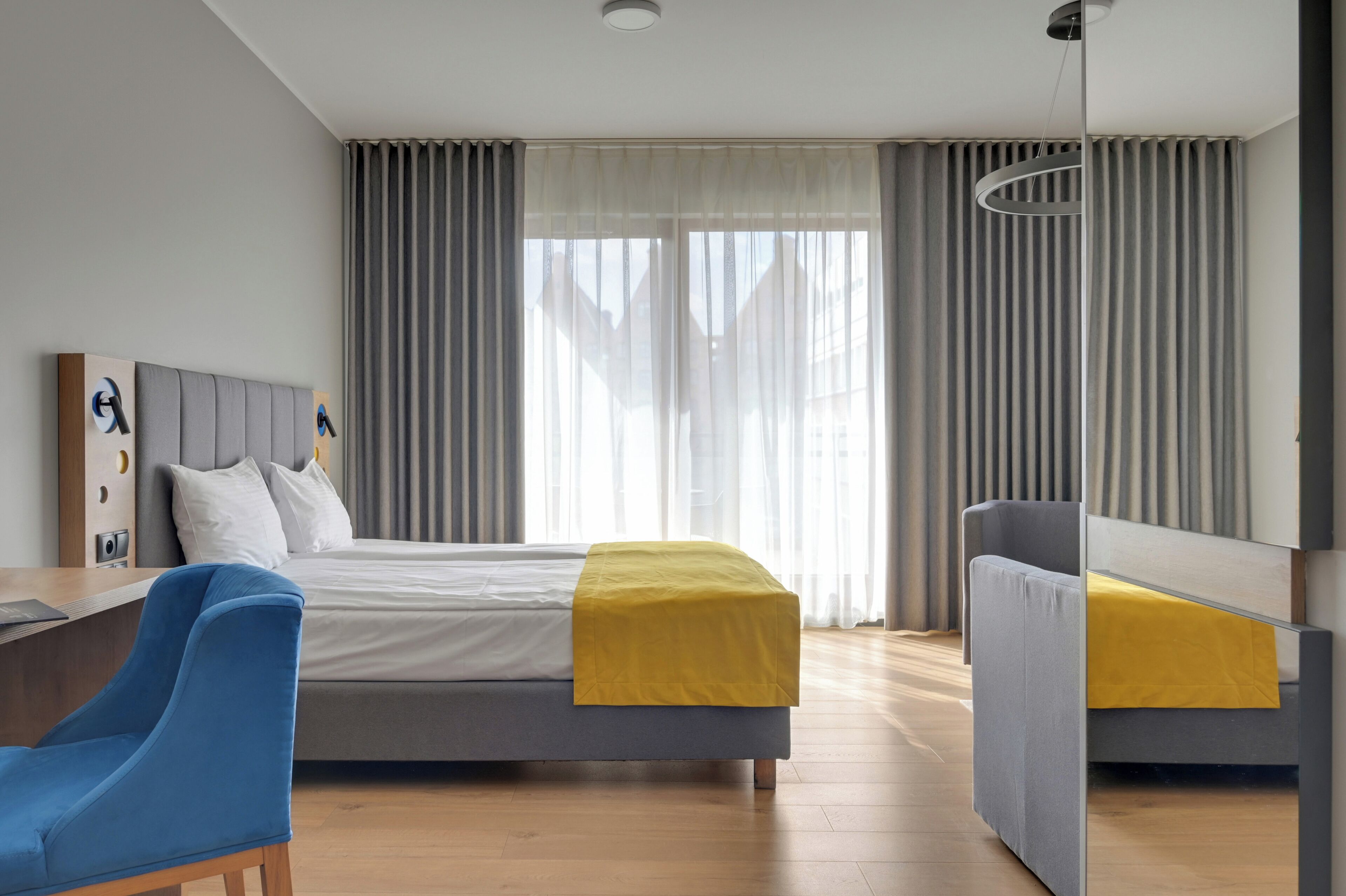 Ein modernes Hotelzimmer mit Bett, Schreibtisch, Sessel und einem großen Fenster mit Vorhängen.