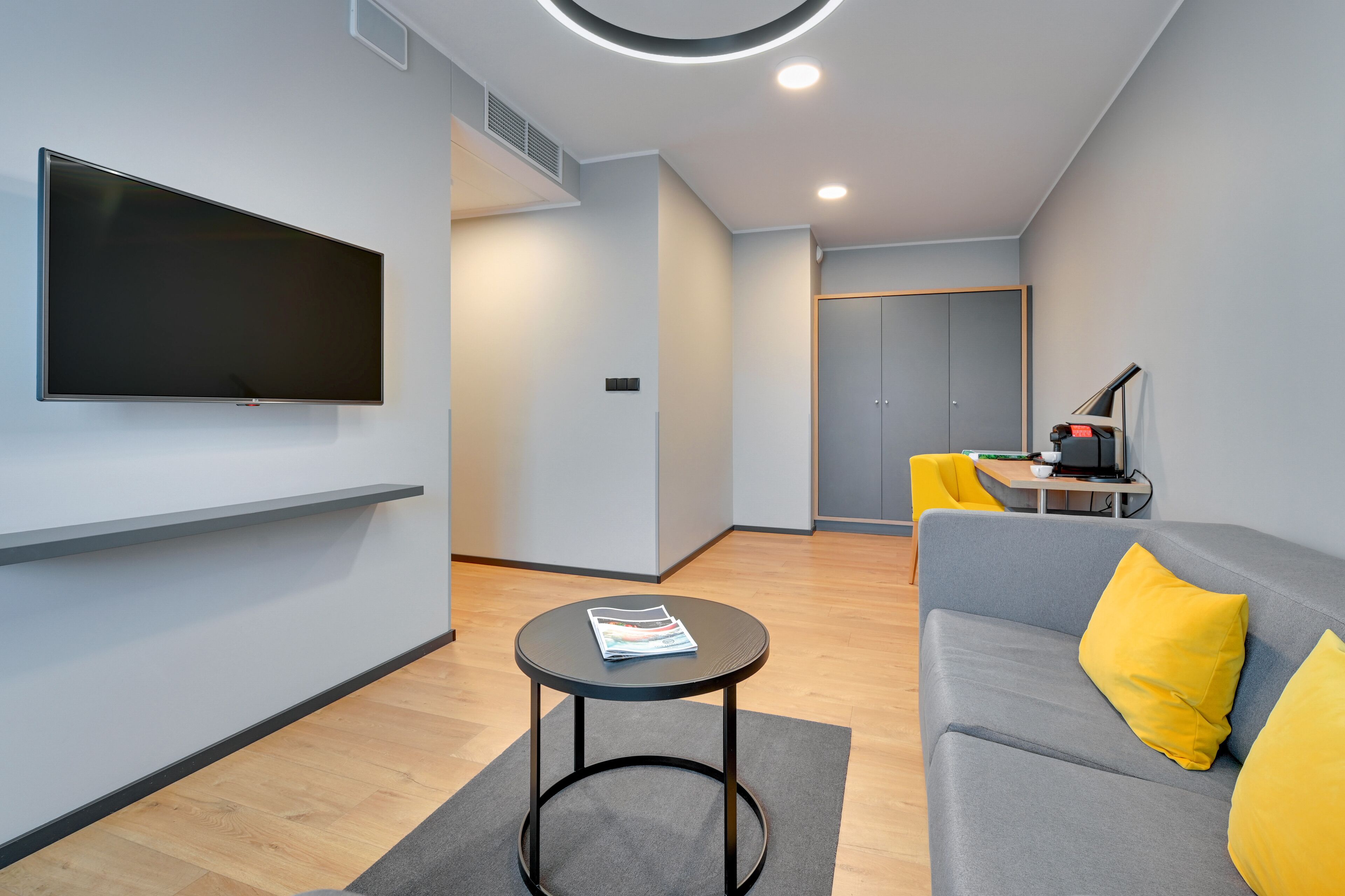 A modern living room with a flat-screen TV, a round table with a magazine, a grey sofa with yellow cushions, and a small desk with a lamp.