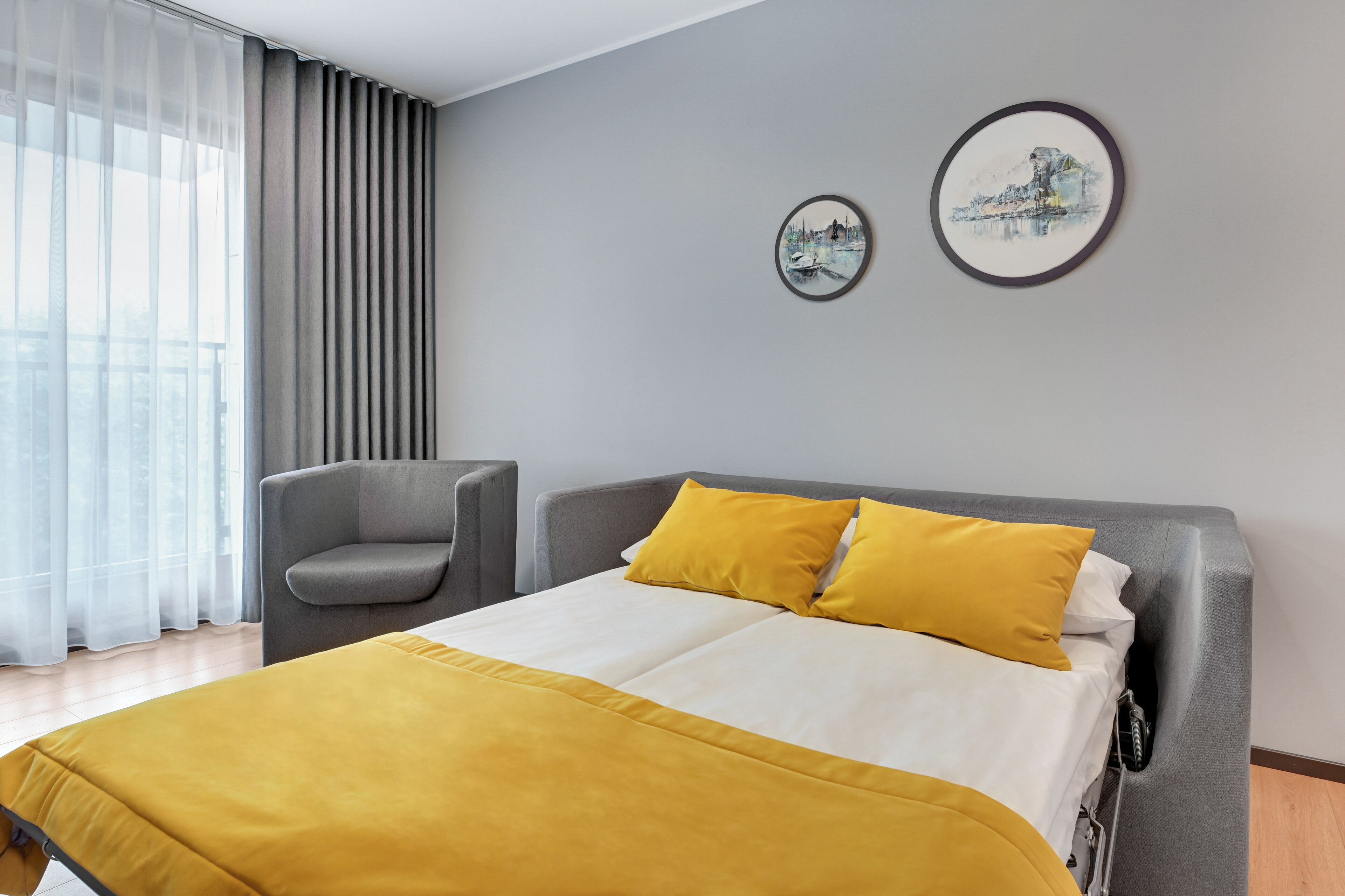 Ein modernes Hotelzimmer mit einem großen Bett, einem grauen Sessel und zwei gerahmten Kunstwerken an der Wand.