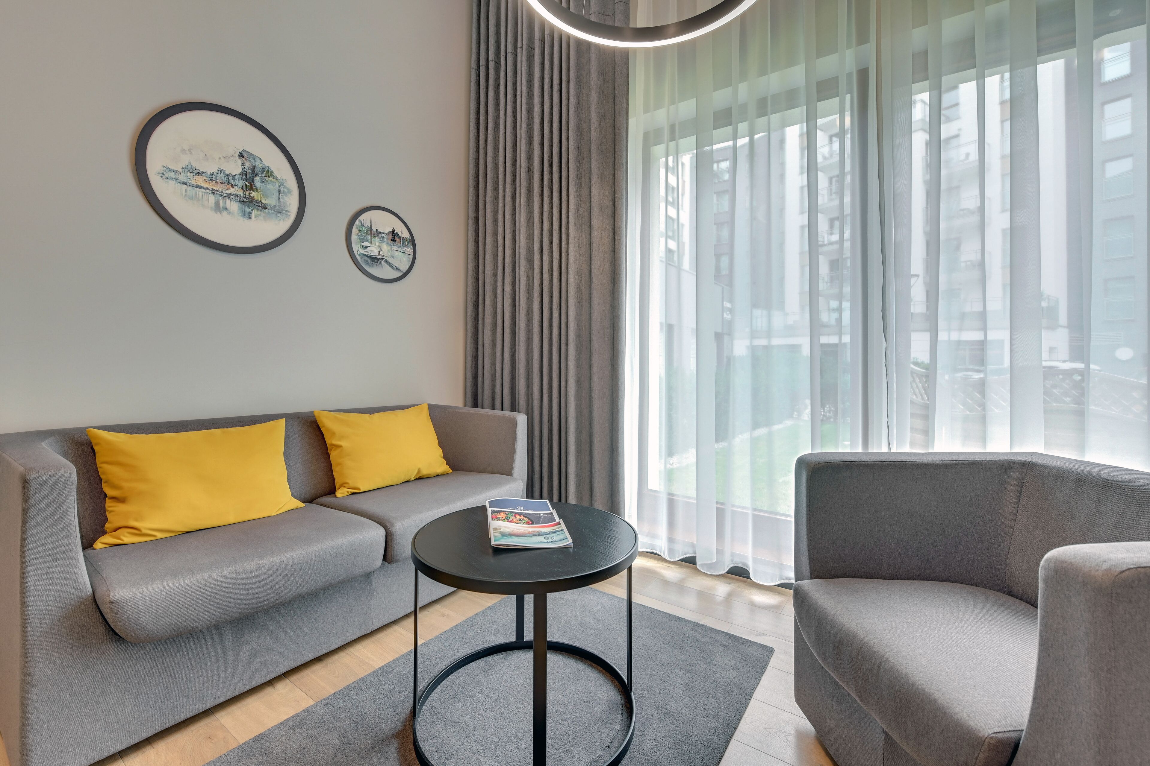 Un salon moderne avec un canapé gris, des coussins jaunes, une table ronde noire et deux tableaux encadrés accrochés au mur.