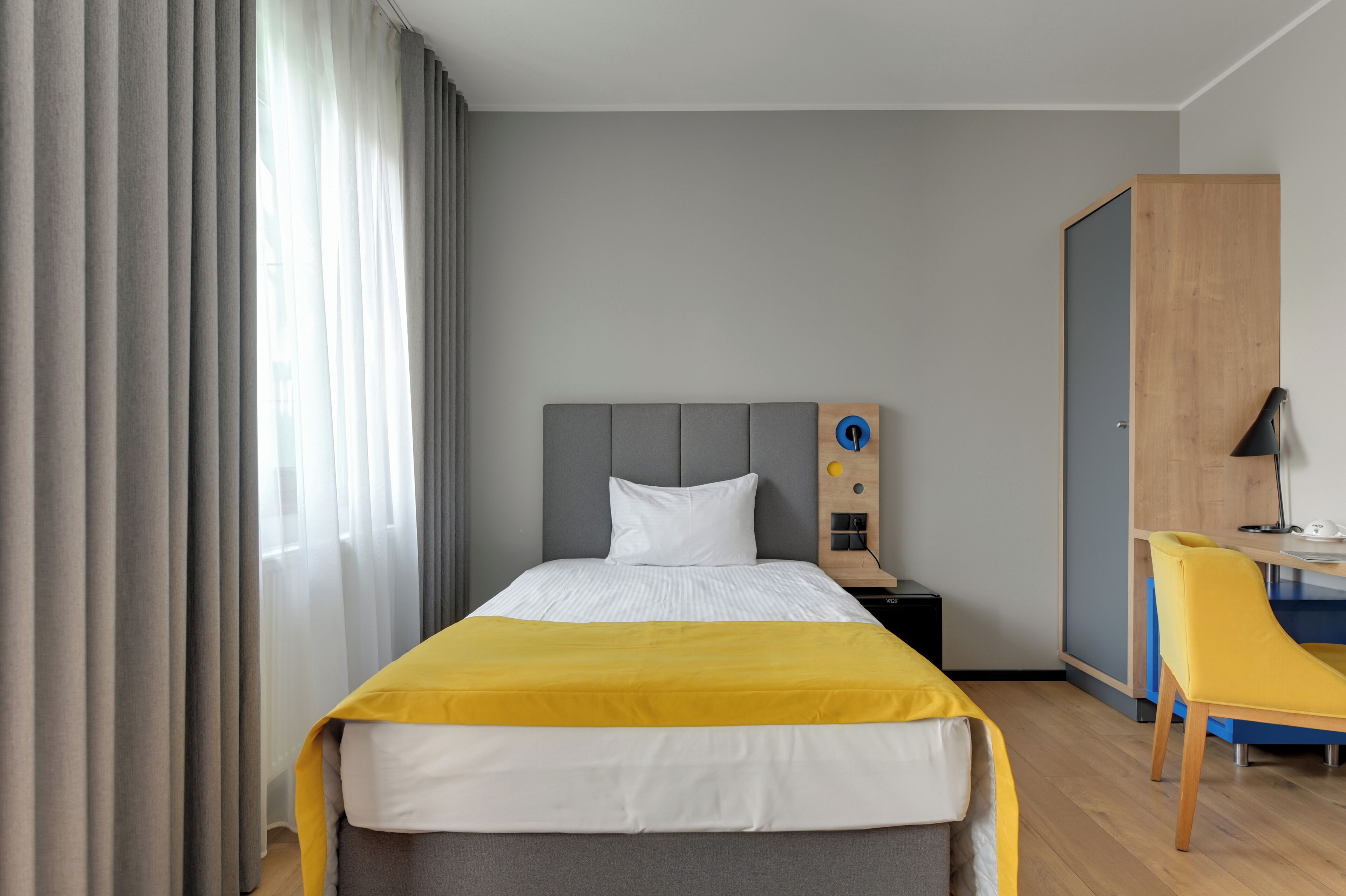A hotel room with a bed, a desk, a chair, a wardrobe, and a window with curtains.