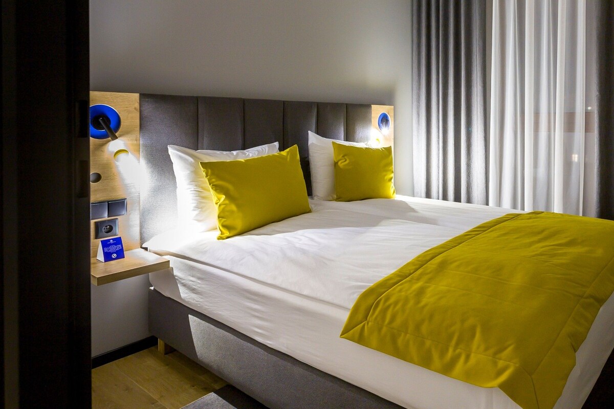 Una habitación de hotel moderna con una cama grande, una mesita de noche y un ventanal con cortinas.