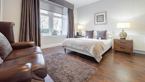 A bedroom with a large bed, a leather armchair, a wooden nightstand, and a window with curtains. - Carmel Apartments Queen Street Residence (Falkirk)