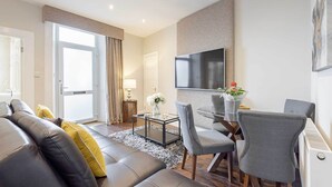 A modern living room with a leather sofa, a dining table with chairs, a flat-screen TV mounted on the wall, and a large window with curtains. - Carmel Apartments Queen Street Residence (Falkirk)