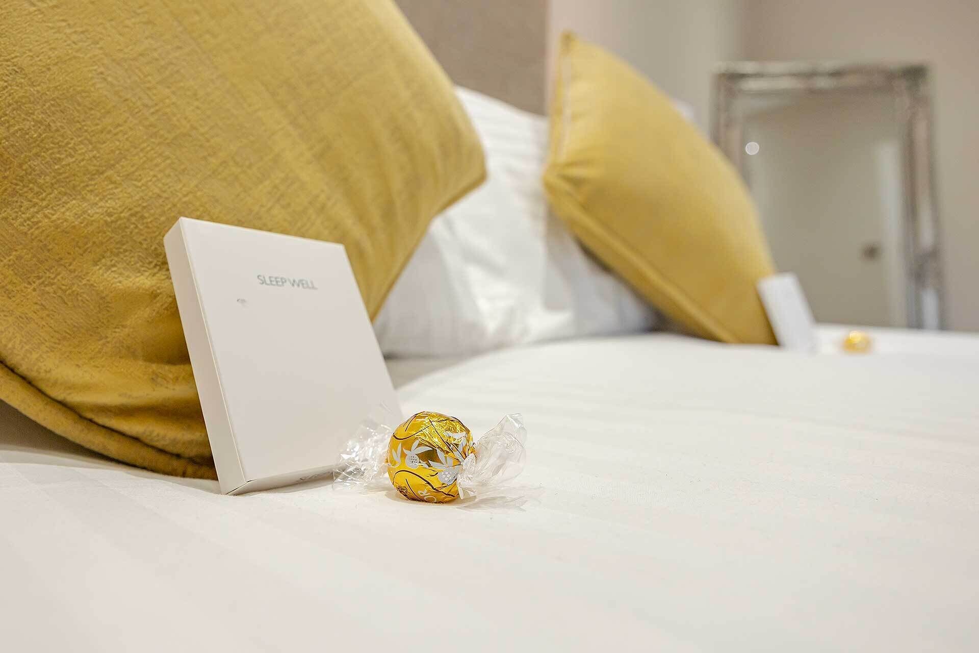 A bed with a white pillow, a white box labeled "SLEEP WELL", and a wrapped chocolate bar on the bed.