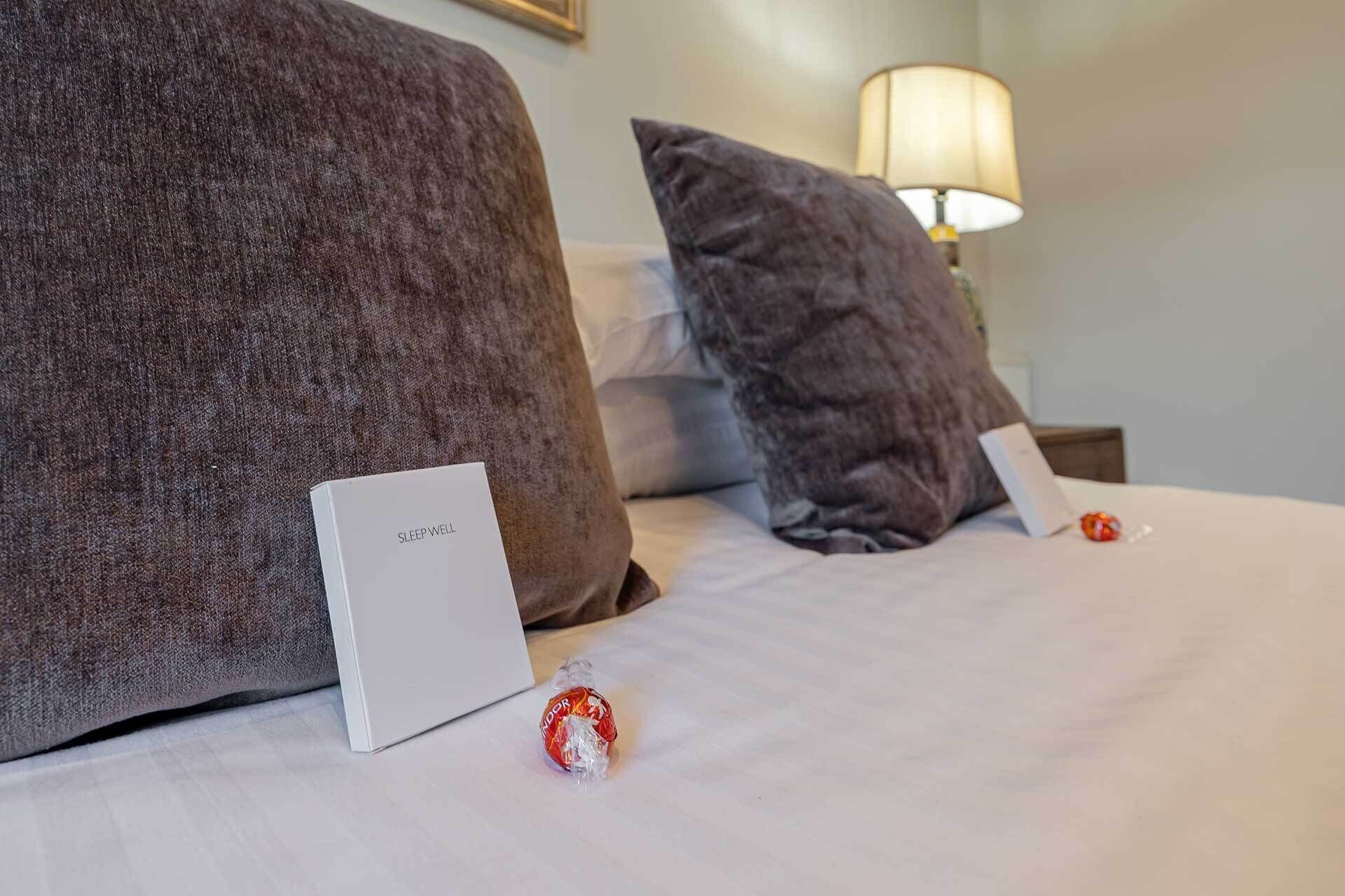Una cama bien hecha con un almohadón blanco y una caja blanca con la etiqueta 'SLEEP WELL' a la izquierda.