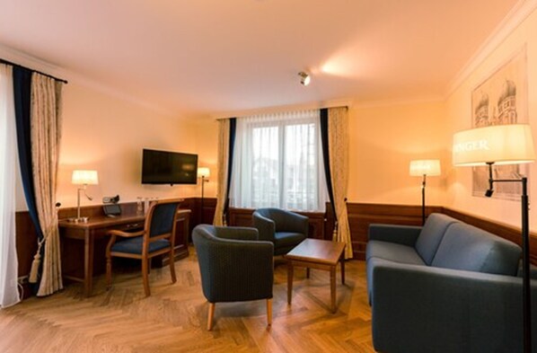 Habitación de hotel con piso de madera, un escritorio con silla, un sofá, un televisor montado en la pared y dos lámparas.