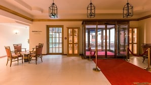 Un vestíbulo de hotel con alfombra roja, sillas de madera, puerta de cristal de entrada y lámparas colgantes.