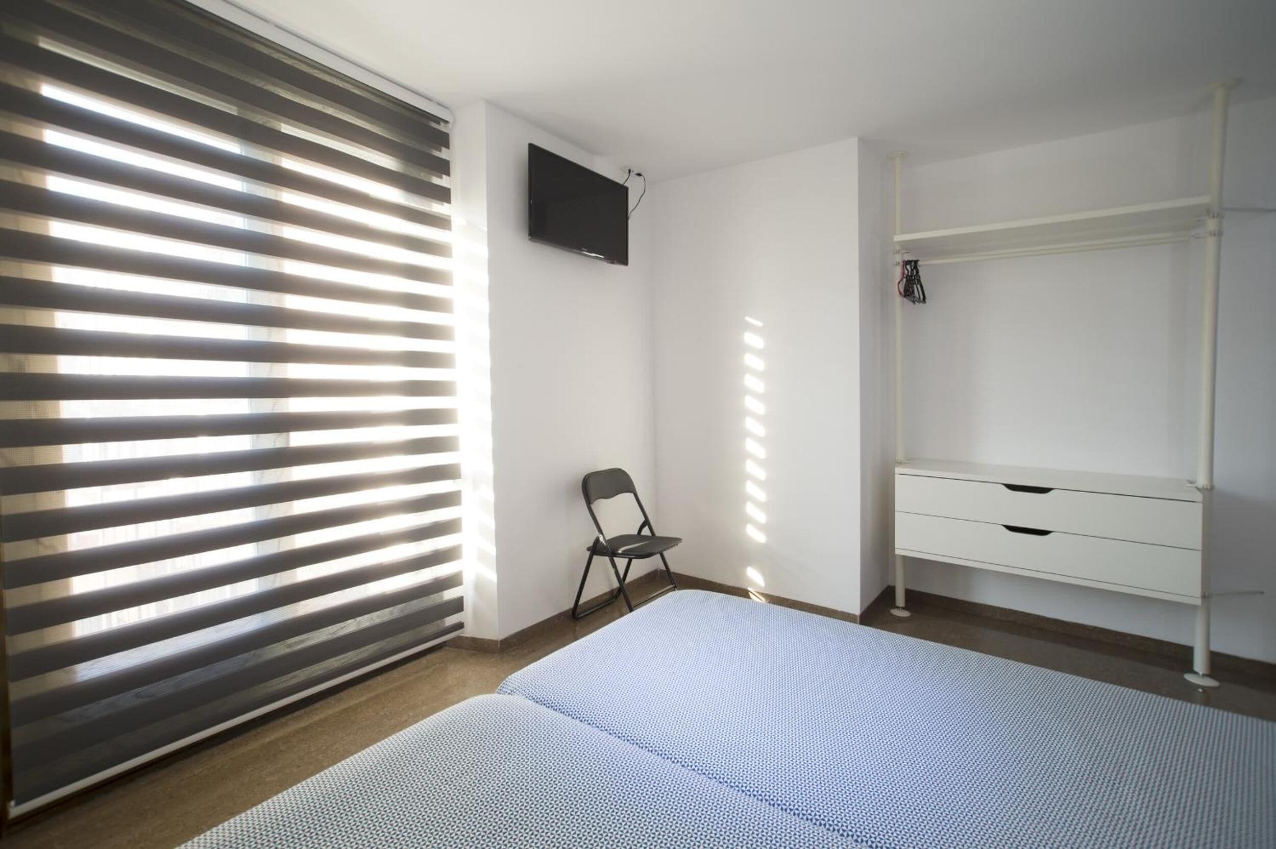 Une chambre à coucher avec un lit, un téléviseur fixé au mur, une chaise et une armoire blanche.