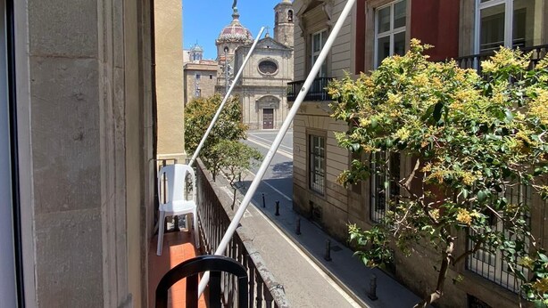 Un balcón con vista a una calle, un edificio con una estatua y un árbol con flores amarillas.