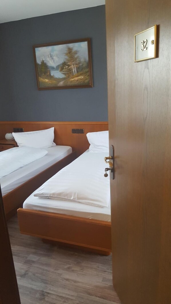 Habitación de hotel con dos camas individuales, cabecero de madera, un cuadro en la pared y una puerta con placa de número.