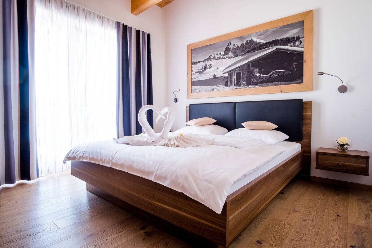 A neatly made bed with white linens and a heart-shaped decoration, a wooden headboard, a nightstand with a vase of flowers, and a framed black and white photograph on the wall.