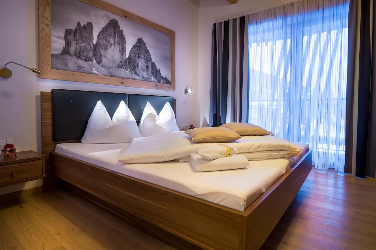 A large bed with a wooden frame, white linens, and multiple pillows. There is a nightstand with a lamp, a painting of mountains on the wall, and a window with curtains.