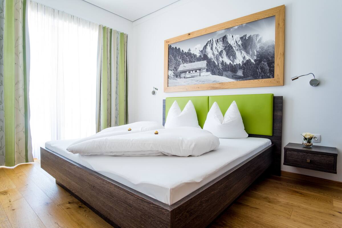 A modern bedroom with a wooden bed frame, white bedding, and a green headboard. A framed photograph of a mountain landscape hangs above the bed.