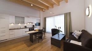 A modern kitchen with white cabinets, a dining area with a wooden table and chairs, a dark brown sofa, and a large window with sheer curtains. - Apartments Dolomit Royal (Sillian)