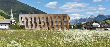 A modern building with a unique wooden facade, surrounded by a field of wildflowers and a church in the background.