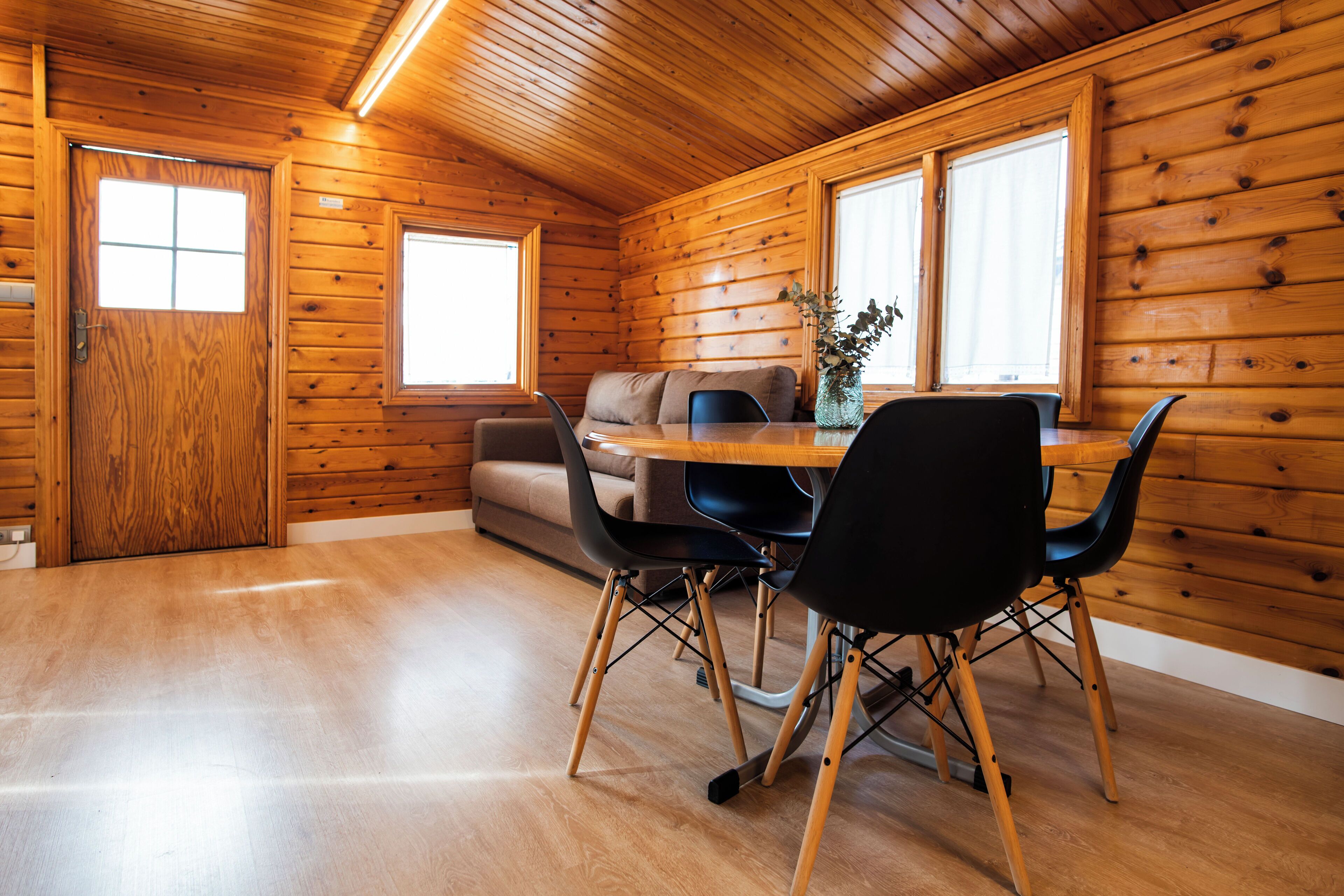 A wooden cabin interior with a dining table, chairs, a sofa, and a window.