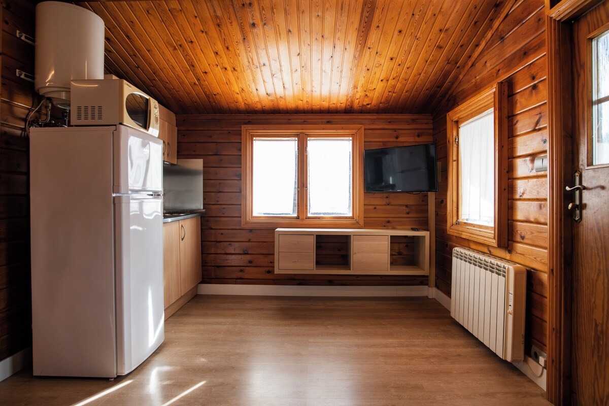 Une cuisine aux murs lambrés, équipée d’un réfrigérateur, d’un four à micro-ondes et d’un four encastré. Deux fenêtres laissent entrer la lumière naturelle et un téléviseur est fixé au mur.