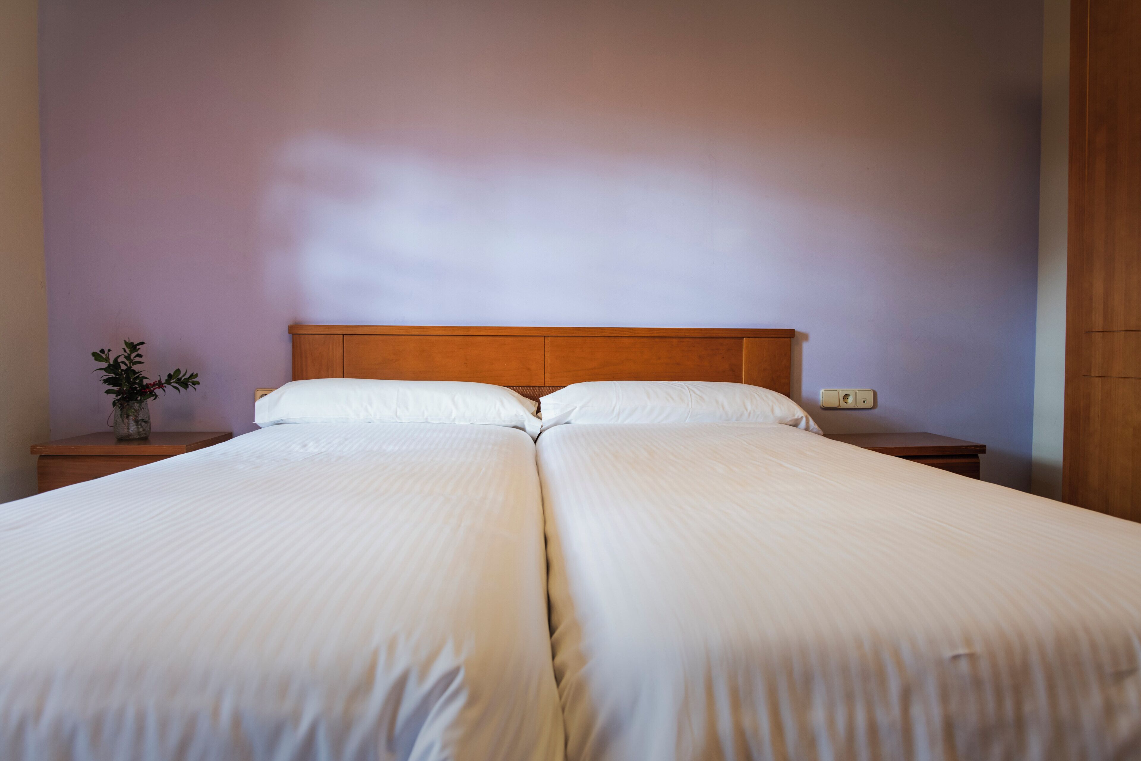 A double bed with white linens, a wooden headboard, and a nightstand with a vase of flowers.