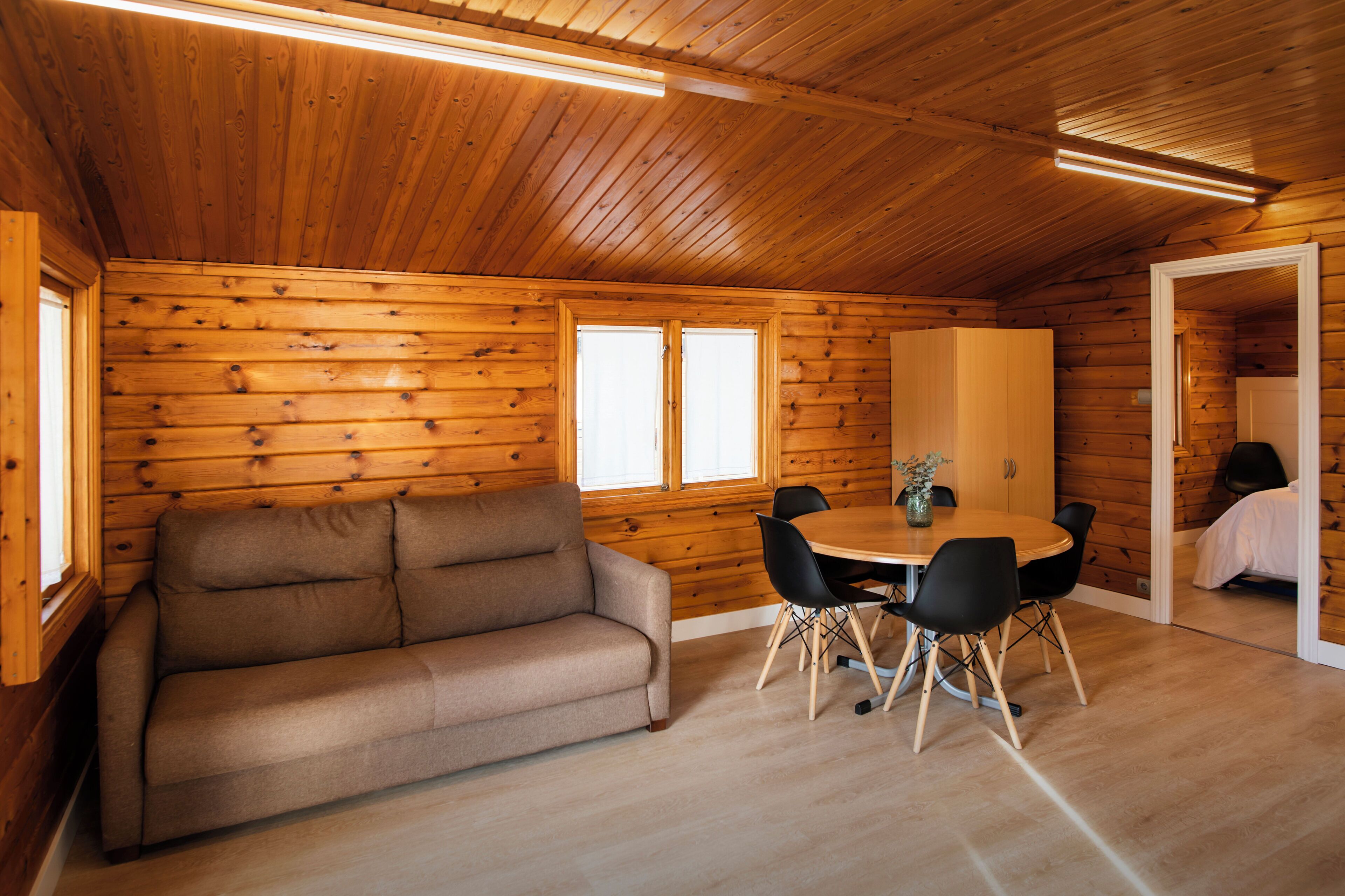 A cozy cabin interior with a wooden ceiling, a sofa, a round dining table with chairs, and a small window.