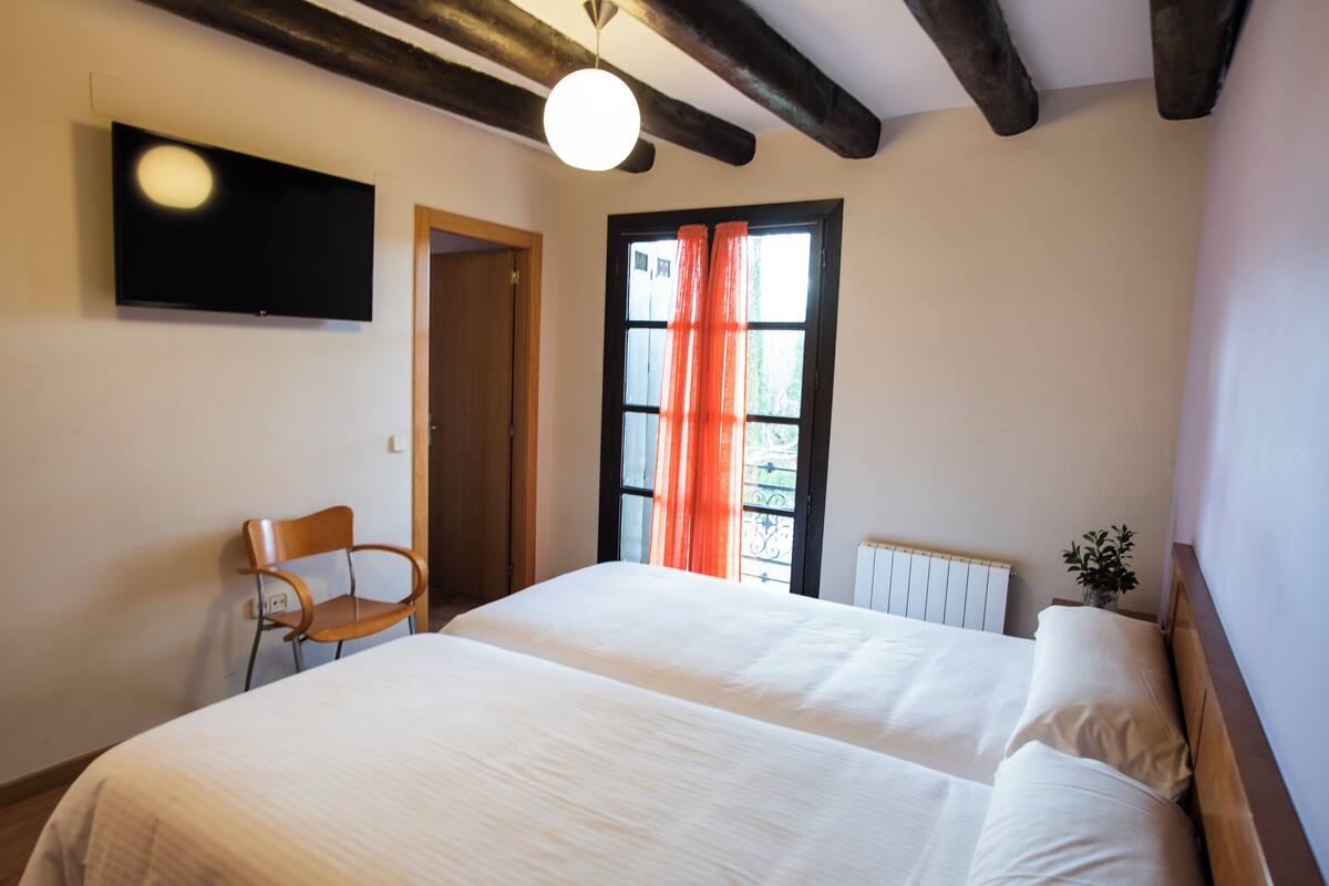 A bedroom with a bed, a chair, a TV, and a window with curtains.