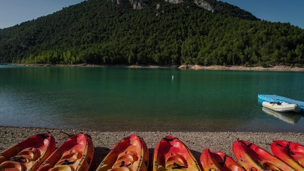 A calm lake with kayaks lined up on the shore and a mountain in the background. - Camping & Bungalows Ligüerre de Cinca (Abizanda)