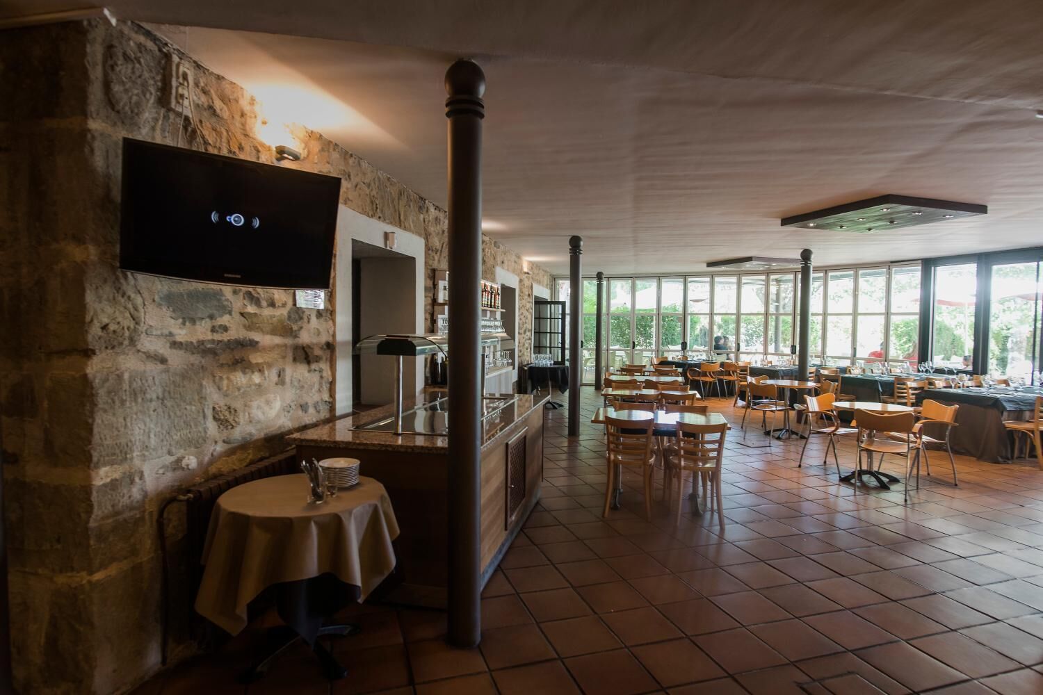 Un restaurante con paredes de piedra, un televisor montado en la pared y una zona de bar con cerramiento de vidrio. Mesas y sillas dispuestas para comer.