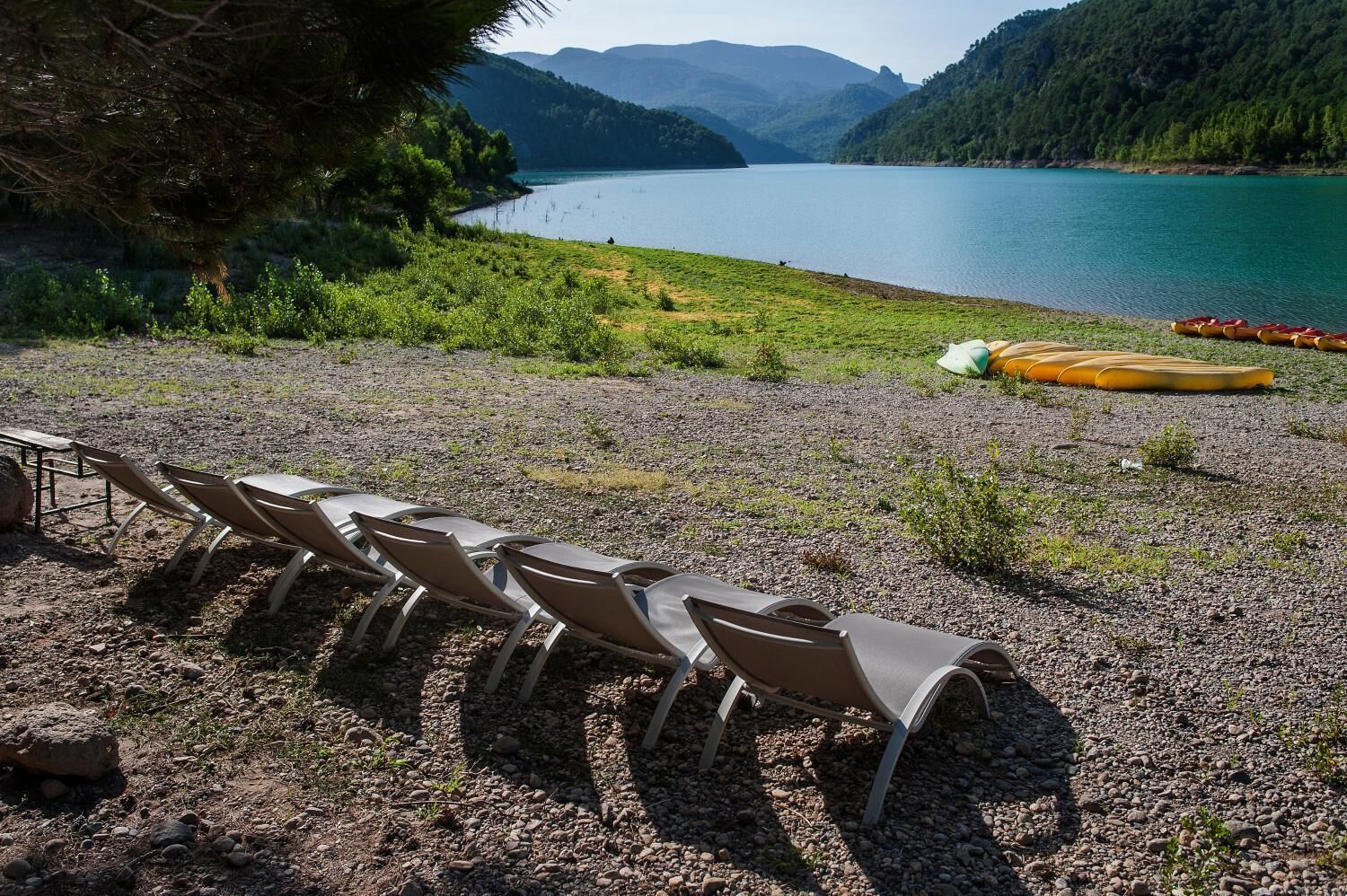 Un espacio de estar junto al lago con sillas y una mesa, rodeado de un terreno rocoso y vistas al lago y las montañas.